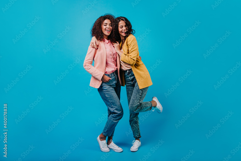 Cheerful latin woman in yellow jacket dancing in studio. Full length portrait of two trendy girls in white shoes. Cheerful latin woman in yellow jacket dancing in studio. Full length portrait of two trendy girls in white shoes.