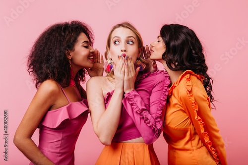 Surprised blonde girl listening friend's secrets. Two brunette ladies sharing rumors with caucasian woman. photo