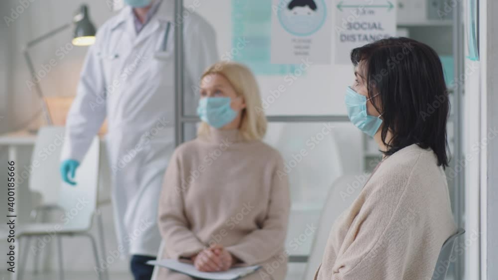 Two women in protective face masks sitting in waiting room in medical office while multiethnic doctor passing by them Two women in protective face masks sitting in waiting room in medical office while multiethnic doctor passing by them