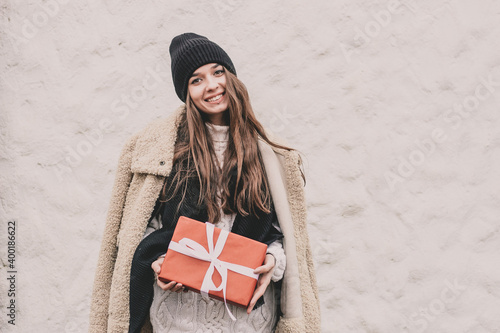 A beautiful young woman of model appearance with long curly hair in a sweater hat and a fur coat holds a Christmas present in her hands on a white background there is a place for an inscription