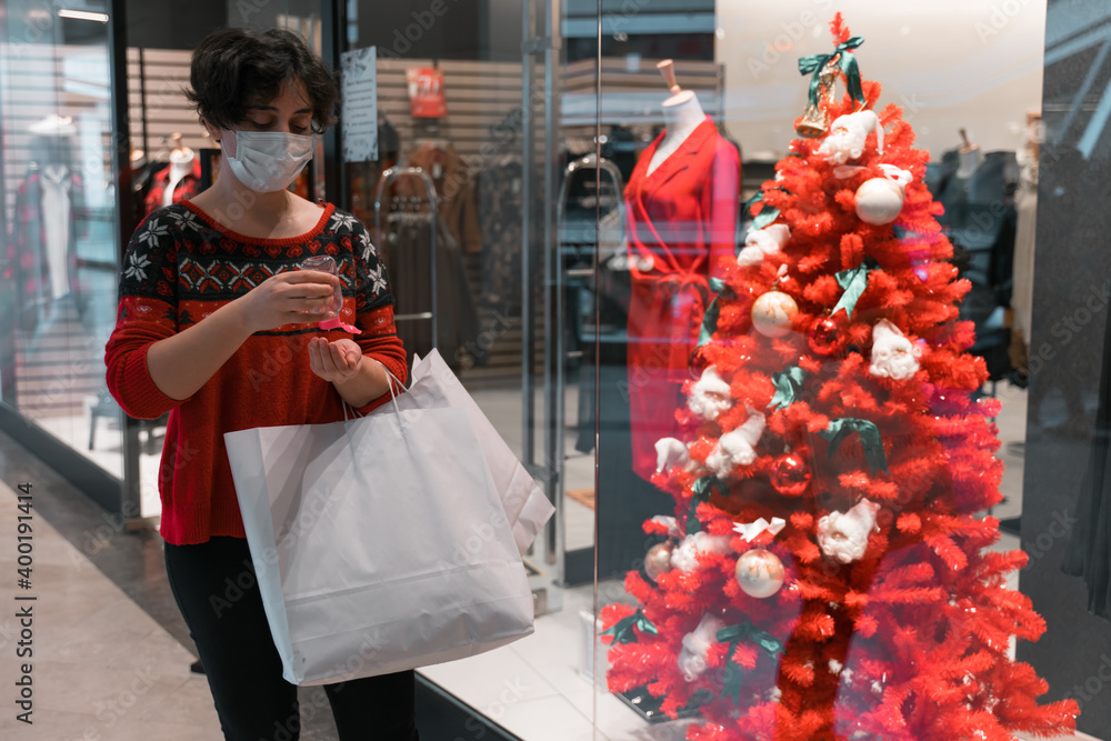 shopping woman wearing medical mask at mall using small portable antibacterial hand sanitizer on hands for christmas or new year. hold shoping bag. ugly christmas sweater. coronavirus pandemic. 2021