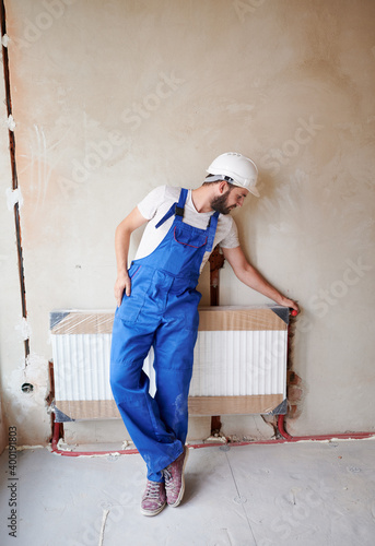 Full length of bearded young man plumber checking radiator while installing heating system in apartment. Male worker wearing blue work overalls and protective helmet, standing near heating battery.