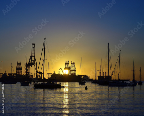 Sunrise from the port, backlit sailboats in the foreground, large ships and cranes in the background and clear blue sky