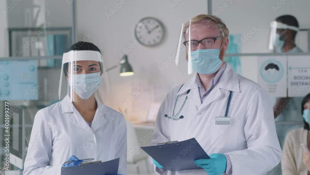 Portrait of Asian female doctor and Caucasian male physician in lab coats, protective mask, screen and gloves holding clipboard and posing for camera in medical office during coronavirus pandemic Portrait of Asian female doctor and Caucasian male physician in lab coats, protective mask, screen and gloves holding clipboard and posing for camera in medical office during coronavirus pandemic