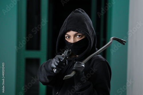 Thief broke into the apartment. House robbery by woman in a black jacket and black mask black gun and crowbar. Burglar in a mask. Thief in a mask trying to break into other people's house.