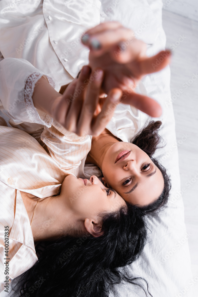 Top view of Hispanic lesbian couple touching hands on blurred foreground while lying on bed at home