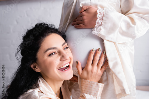 Cheerful Hispanic woman listening belly of pregnant partner at home