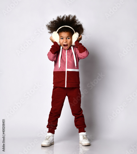 African American girl stands in warm headphones and sportswear and shouts loudly, covering her ears with her hands. Child poses on a gray background. Concept of children's emotions and sports clothes.