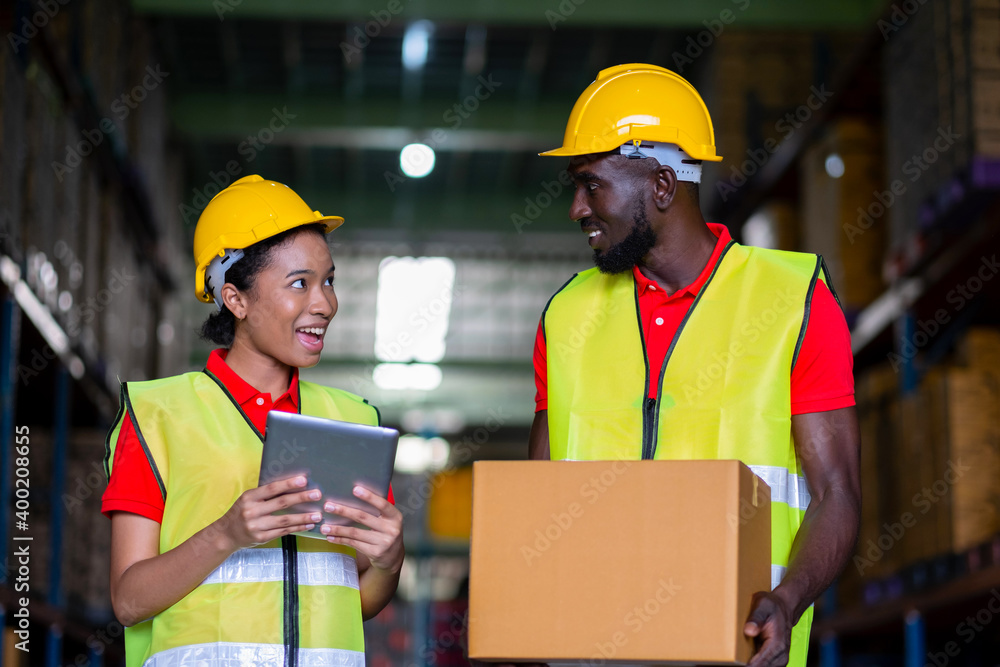 Woman worker using a tablet to check stock online and a man holding a parcel in the automotive parts warehouse center. Both engineers people wear a safety helmet. In background shelves with goods Woman worker using a tablet to check stock online and a man holding a parcel in the automotive parts warehouse center. Both engineers people wear a safety helmet. In background shelves with goods