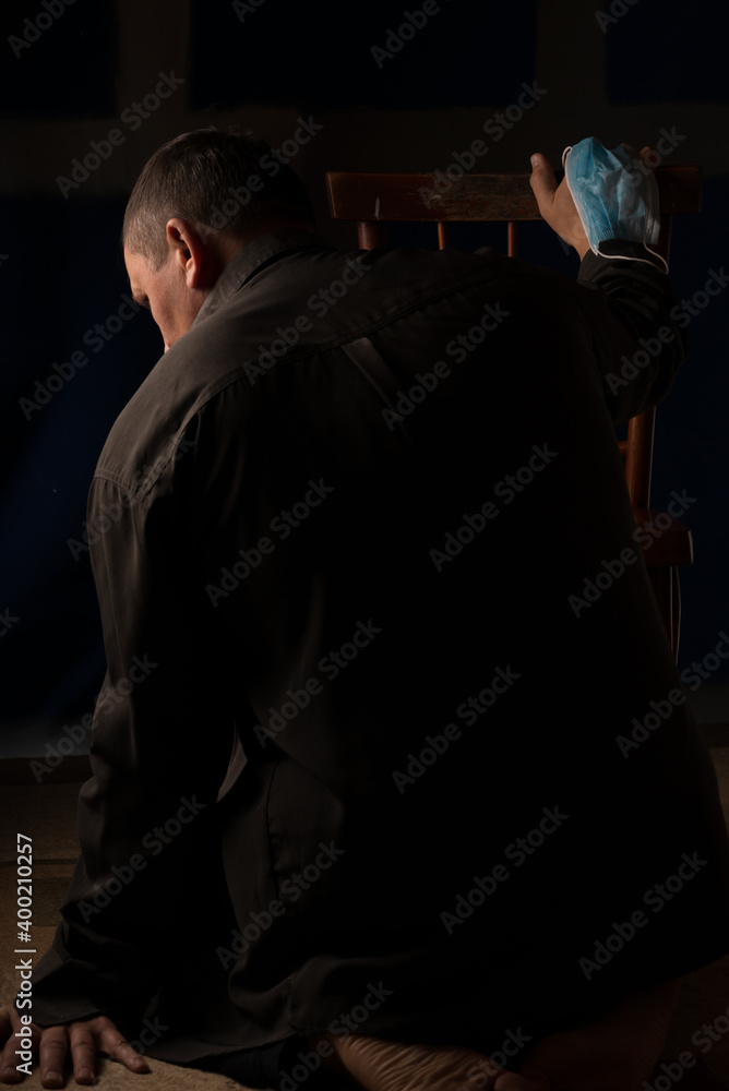 The man sits with his back in a dark room. Leans on a chair. Medical mask. A bright emotional state. The man sits with his back in a dark room. Leans on a chair. Medical mask. A bright emotional state.