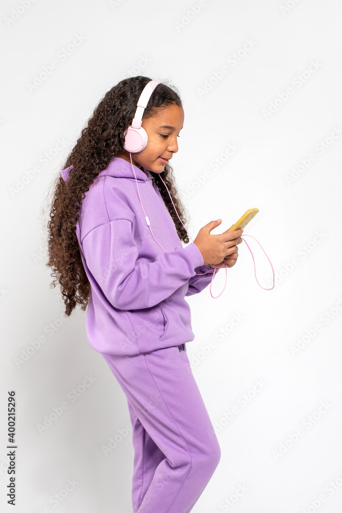 Young dark skinned African girl with dark skin wearing lilac tracksuit stands or walks in pink headphones and with a phone. Lifestyle of children and adults. Young dark skinned African girl with dark skin wearing lilac tracksuit stands or walks in pink headphones and with a phone. Lifestyle of children and adults.