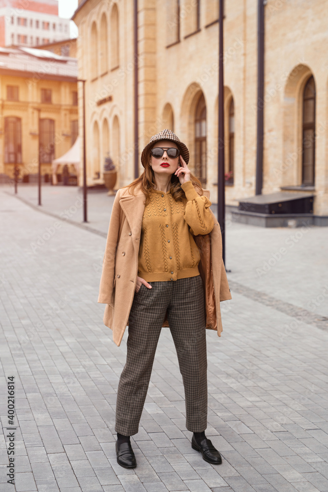 Full height self confident girl in an autumn beige coat and sunglasses, plaid panama hat standing posing on the street with coat on her shoulders. Retro toned photo. 