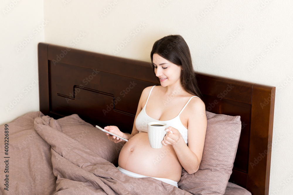 pregnant young woman sitting on bed and drinking coffee and texting message at home. maternity concept pregnant young woman sitting on bed and drinking coffee and texting message at home. maternity concept