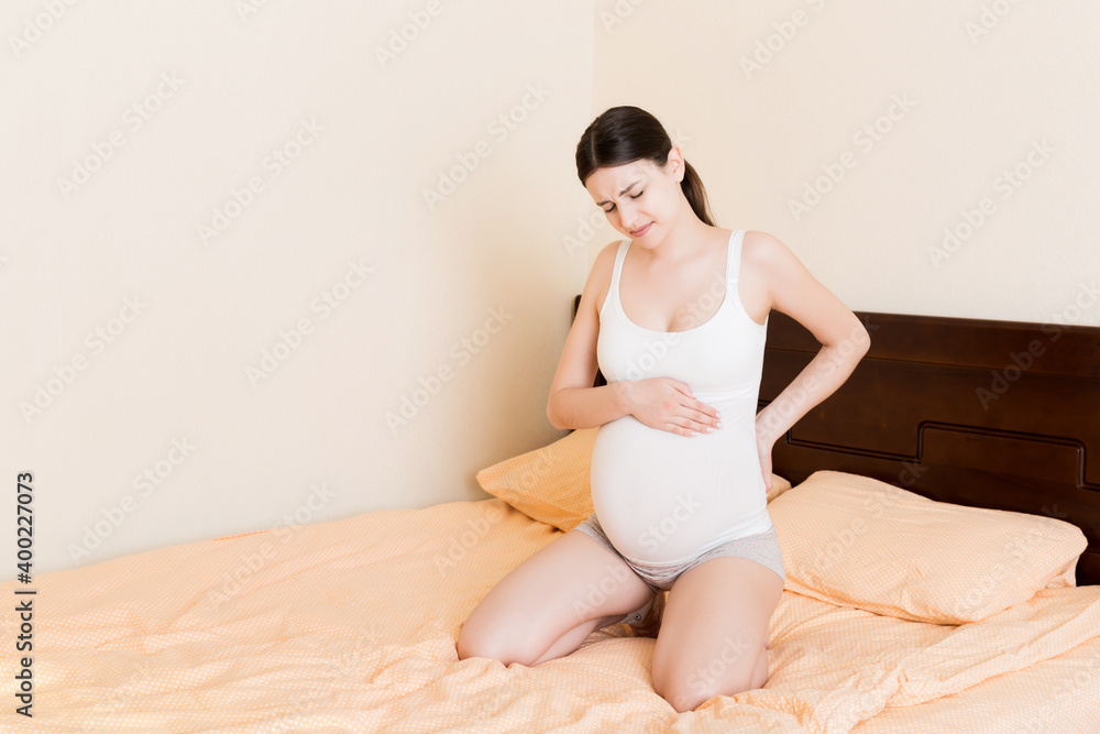beautiful pregnant woman touching her tummy and keeping one hand on her back at home on bed beautiful pregnant woman touching her tummy and keeping one hand on her back at home on bed