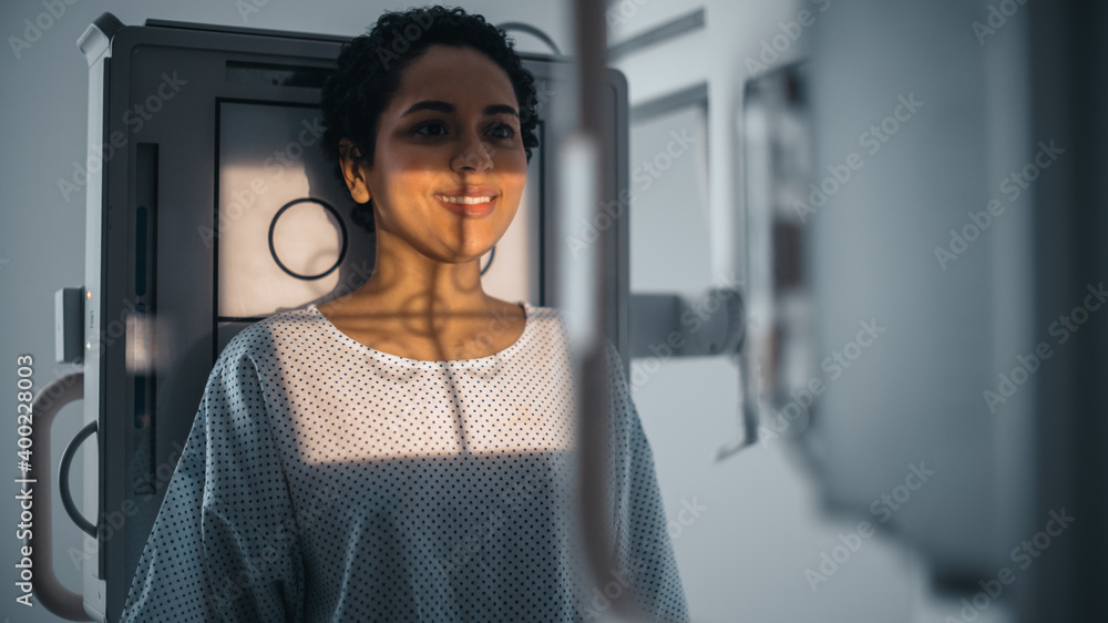 Hospital Radiology Room: Portrait of Beautiful Latin Woman Standing next to X-Ray Machine While it Scans Chest, Back, Lungs. Modern High-Tech Laboratory with Advanced Technical Equipment. She Smiles.