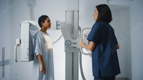 Hospital Radiology Room  Beautiful Latin Woman Standing while Professional Female Radiologist Explains Procedure  Adjusts X-Ray Machine. Scanning Chest  Heart  Lungs. Mammogram Cancer Screening