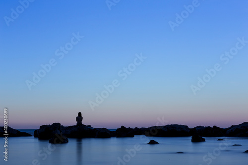 Young woman silhouette sitting on some rocks surrounded by water in an incredible sunset.