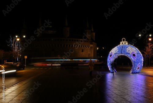 Night view of street decorated with bright illuminated Christmas tree bauble, in background colorful trails of tram lights and historical building called Barbakan, Krakow, Poland photo