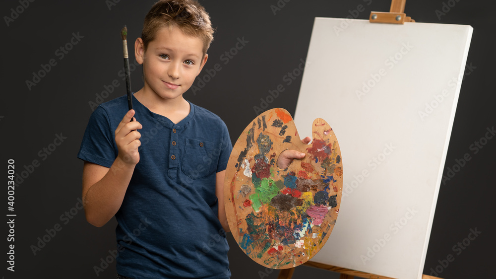 Fototapeta premium Holding a brush and palette with paints 10 year old boy in blue t-shirt looks at camera on the background of an easel with canvas. School of arts concept. Art concept. 