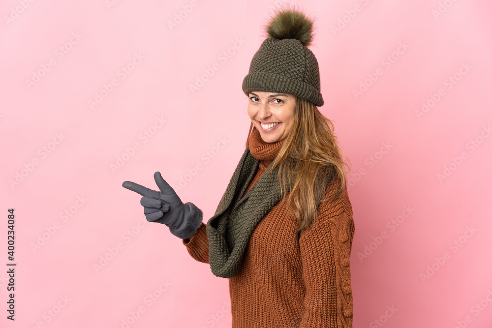 Middle age woman with winter hat isolated on pink background pointing finger to the side Middle age woman with winter hat isolated on pink background pointing finger to the side