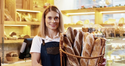 Close up portrait of happy young Caucasian pretty woman seller in apron holding basket with fresh baked bread in hands standing in small bakehouse, looking at camera and smiling alone. Work concept