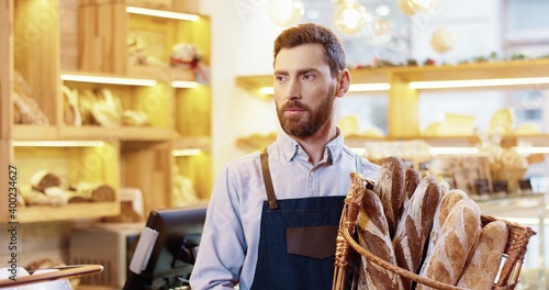 Close up portrait of happy young Caucasian handsome bearded man seller in apron holding basket with fresh baked bread standing in bakehouse, looking at camera and smiling alone. Worker concept
