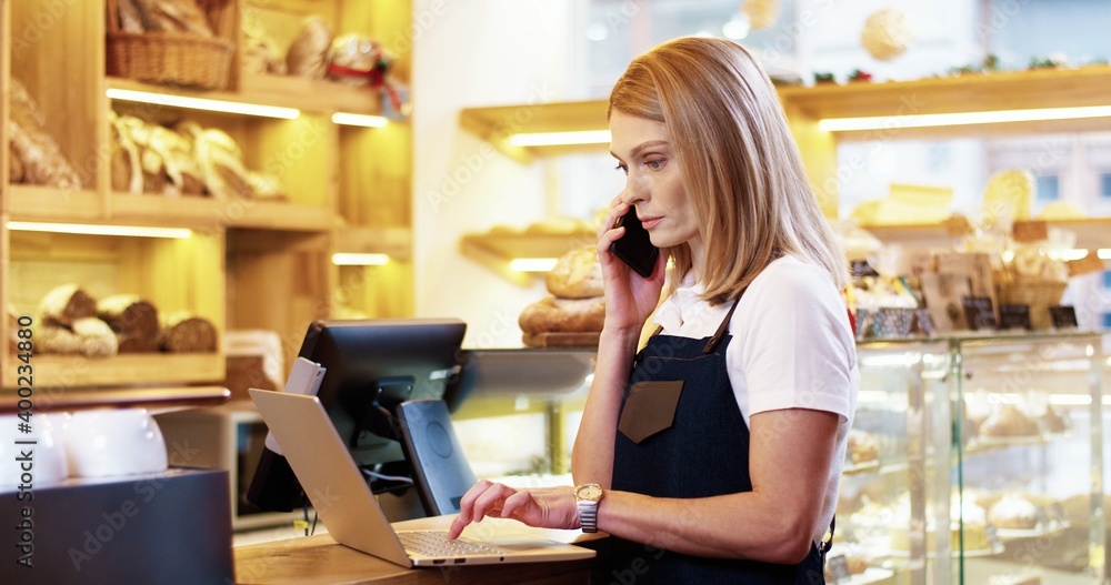 Close up of beautiful young Caucasian woman seller in apron standing in bakery, tapping and typing on laptop talking to male colleague working on background. Bakehouse concept. Small business Close up of beautiful young Caucasian woman seller in apron standing in bakery, tapping and typing on laptop talking to male colleague working on background. Bakehouse concept. Small business