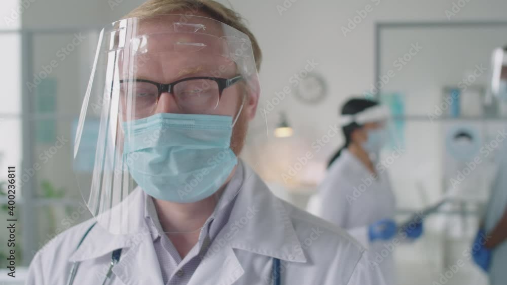 Chest up tracking shot of male doctor in protective face screen, lab coat and medical mask looking at camera while working in hospital during coronavirus pandemic Chest up tracking shot of male doctor in protective face screen, lab coat and medical mask looking at camera while working in hospital during coronavirus pandemic