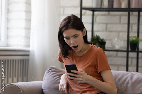 Out of juice. Shocked millennial woman sitting on sofa at home seeing no charge or wifi signal at moment of important call. Female student feeling mad of anger because her cell is being broken hacked