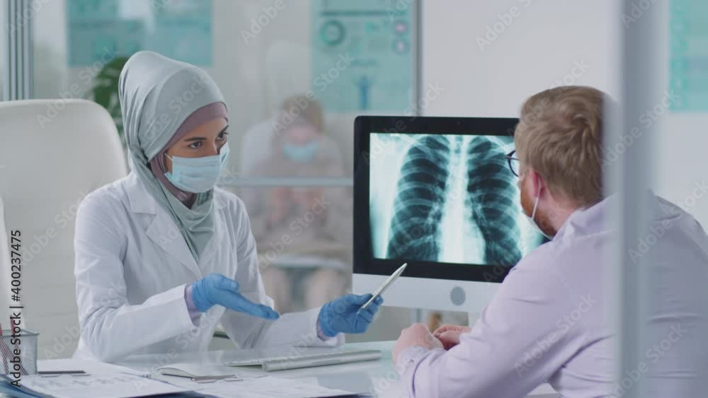 Muslim female doctor in hijab and disposable face mask showing chest x-ray on computer to male patient while giving consultation in medical office during covid-19 pandemic Muslim female doctor in hijab and disposable face mask showing chest x-ray on computer to male patient while giving consultation in medical office during covid-19 pandemic