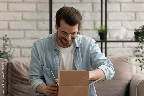 What is inside. Curious millennial male sitting on couch at home opening small cardboard box received by mail. Interested young man unpacking shipment delivered to addressee by express courier service photo