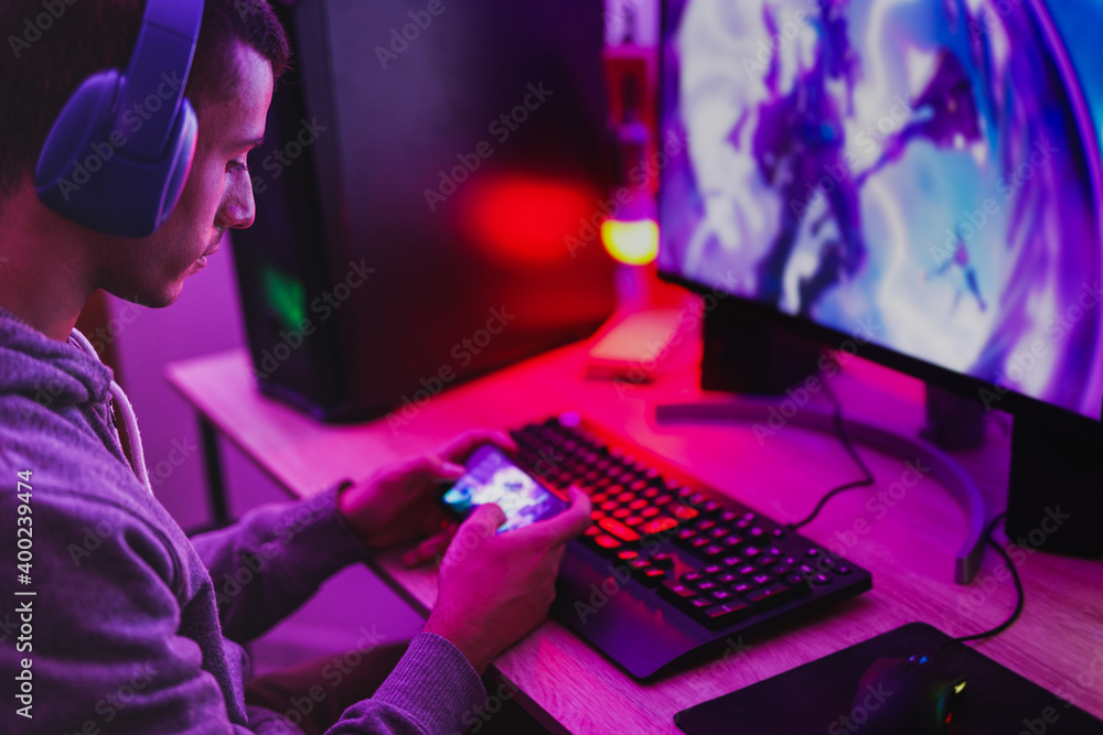 Young boy wearing headset and play video games with smartphone online in front of computer - Home isolated for coronavirus outbreak Young boy wearing headset and play video games with smartphone online in front of computer - Home isolated for coronavirus outbreak