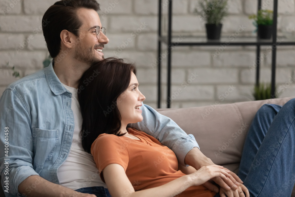 Visualizing dreams. Happy serene young wife resting on sofa reclining back to dear husband feeling support care in his arms. Loving millennial couple dreaming together smiling looking at distance Visualizing dreams. Happy serene young wife resting on sofa reclining back to dear husband feeling support care in his arms. Loving millennial couple dreaming together smiling looking at distance