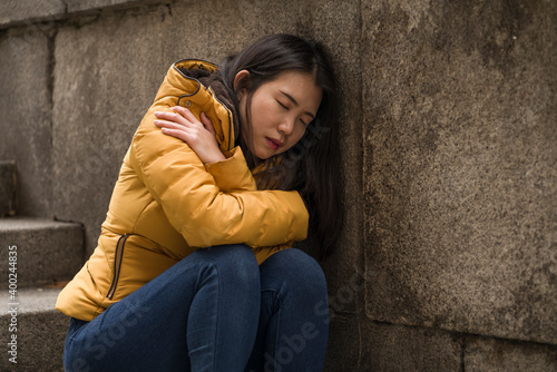 young attractive sad and depressed Chinese woman in winter jacket sitting outdoors on street corner staircase suffering depression problem feeling helpless