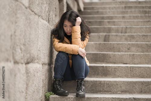 dramatic lifestyle portrait of young attractive sad and depressed Japanese woman in winter jacket sitting outdoors on street corner staircase suffering depression problem feeling helpless