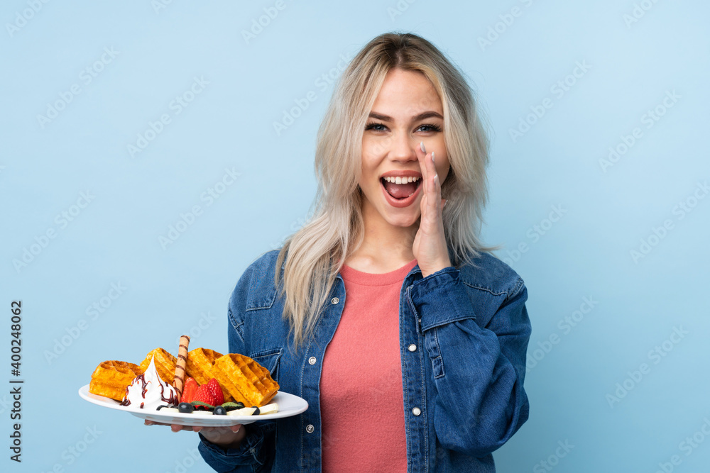 Teenager girl holding waffles over isolated blue background shouting with mouth wide open