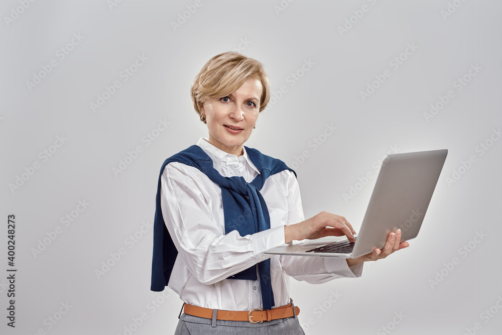 Portrait of elegant middle aged caucasian woman wearing business attire looking at camera while holding and using laptop, posing isolated over grey background Portrait of elegant middle aged caucasian woman wearing business attire looking at camera while holding and using laptop, posing isolated over grey background