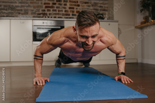 A happy muscular man with a beard is doing pushups on a blue yoga mat in his apartment in the evening. An athletic guy with tattoos on his forearms is doing a chest and triceps workout at home.
