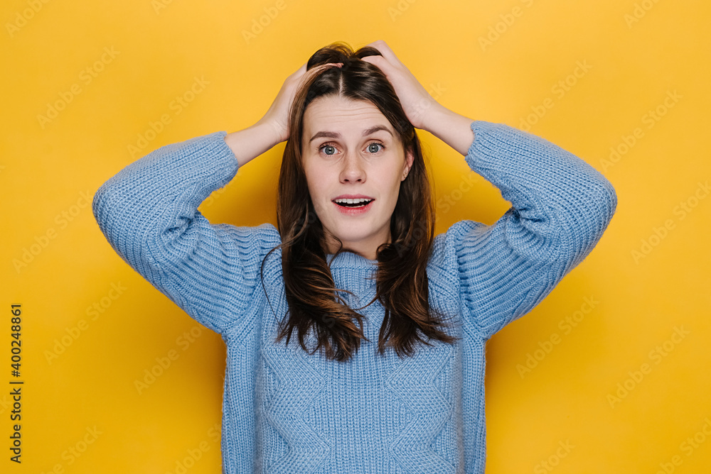 Portrait of hysterical young woman expression with her hands on head, stressed looking at camera, dressed in blue sweater, isolated on yellow studio background. People emotional lifestyle concept Portrait of hysterical young woman expression with her hands on head, stressed looking at camera, dressed in blue sweater, isolated on yellow studio background. People emotional lifestyle concept