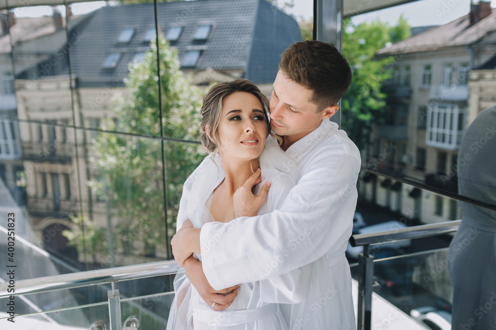 A handsome groom and a sweet bride in white dressing gowns tenderly embrace against the background of a mirrored wall in an expensive hotel. Morning of the newlyweds. Wedding portrait. A handsome groom and a sweet bride in white dressing gowns tenderly embrace against the background of a mirrored wall in an expensive hotel. Morning of the newlyweds. Wedding portrait.