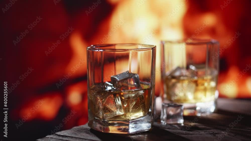 A glass of whiskey with ice is on a wooden table. In the back, a bright fire burns in the fireplace. A glass of whiskey with ice is on a wooden table. In the back, a bright fire burns in the fireplace.