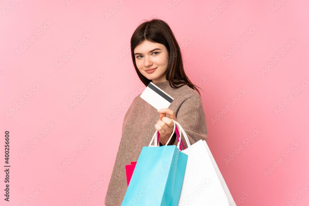 Young Ukrainian teenager girl over isolated pink background holding shopping bags and a credit card