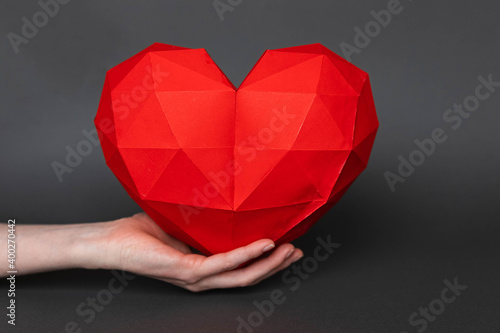 Two female hands  holding a red polygonal paper in the shape of a heart  on a gray background. Red polygonal paper heart for Valentine s Day or any other love invitations.