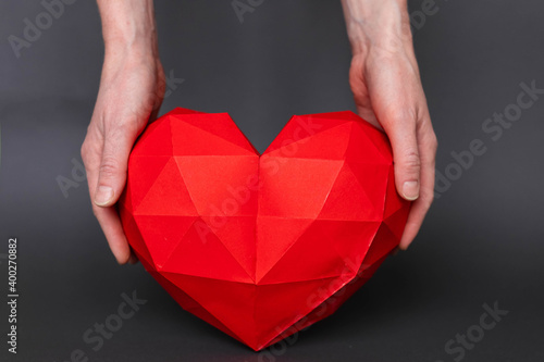 Two female hands  holding a red polygonal paper in the shape of a heart  on a gray background. Red polygonal paper heart for Valentine s Day or any other love invitations.