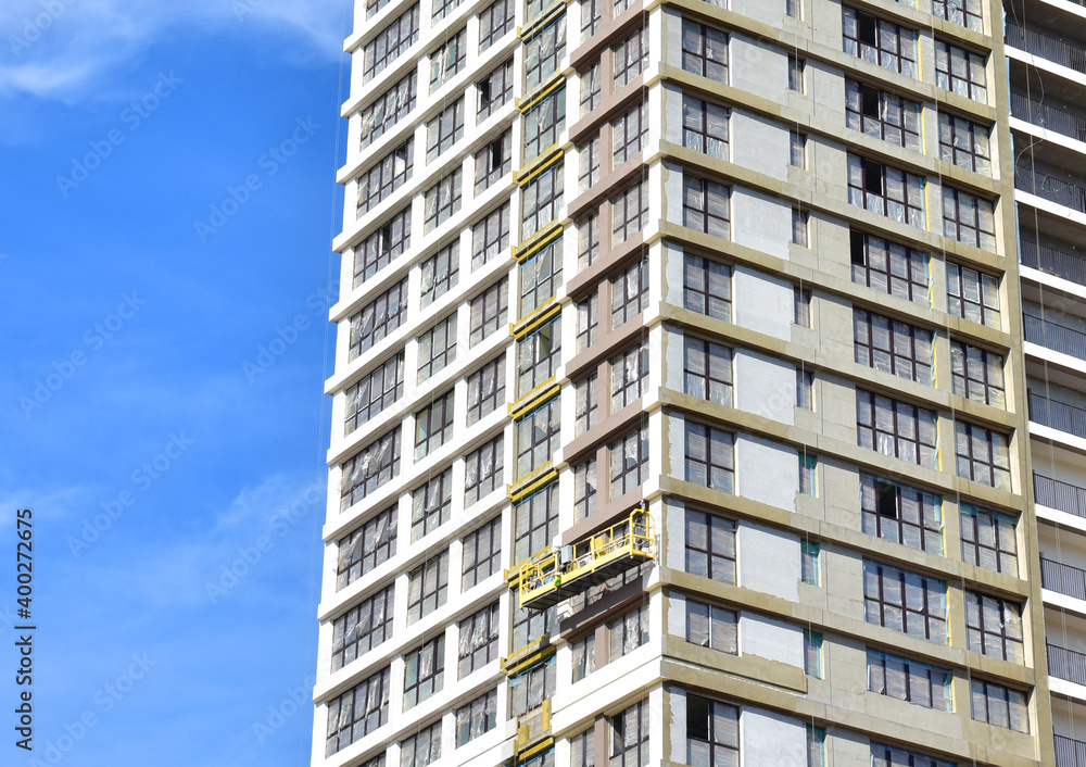 View of the facade of a new multi-storey building under construction. Suspended cradle on the facade of the skyscraper. Mobile scaffolding platform for construction work at height View of the facade of a new multi-storey building under construction. Suspended cradle on the facade of the skyscraper. Mobile scaffolding platform for construction work at height