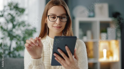 Adorable dark-haired woman in casual outfit and eyewear looking at digital tablet while sitting at table. Young brunette using modern gagdet during free time at home. photo