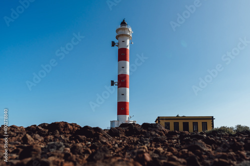 An old lighthouse with white and red stripes on the shore of a rocky island. Clear sunny blue sky.