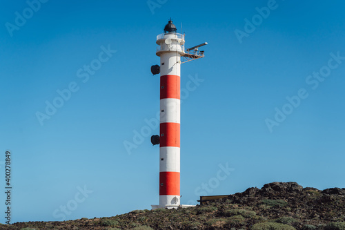 An old lighthouse with white and red stripes on the shore of a rocky island. Clear sunny blue sky.
