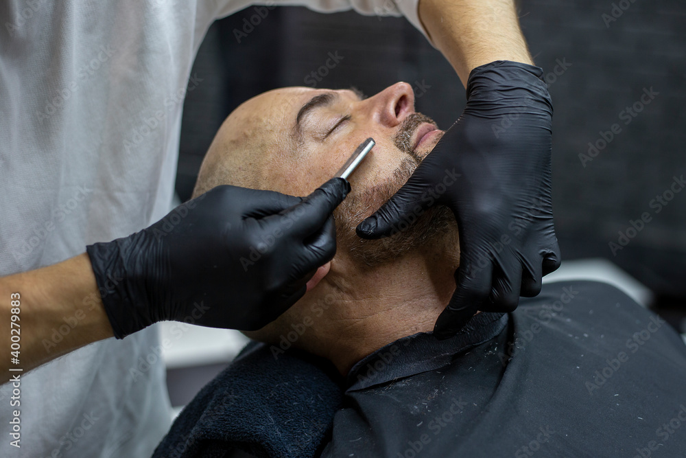Hands of a barber in black gloves shave the beard of a client with a straight razor in the barber shop