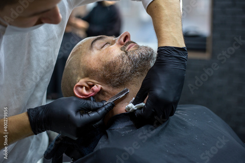 Barber in black gloves shaves the beard of a client with a straight razor in the barber shop